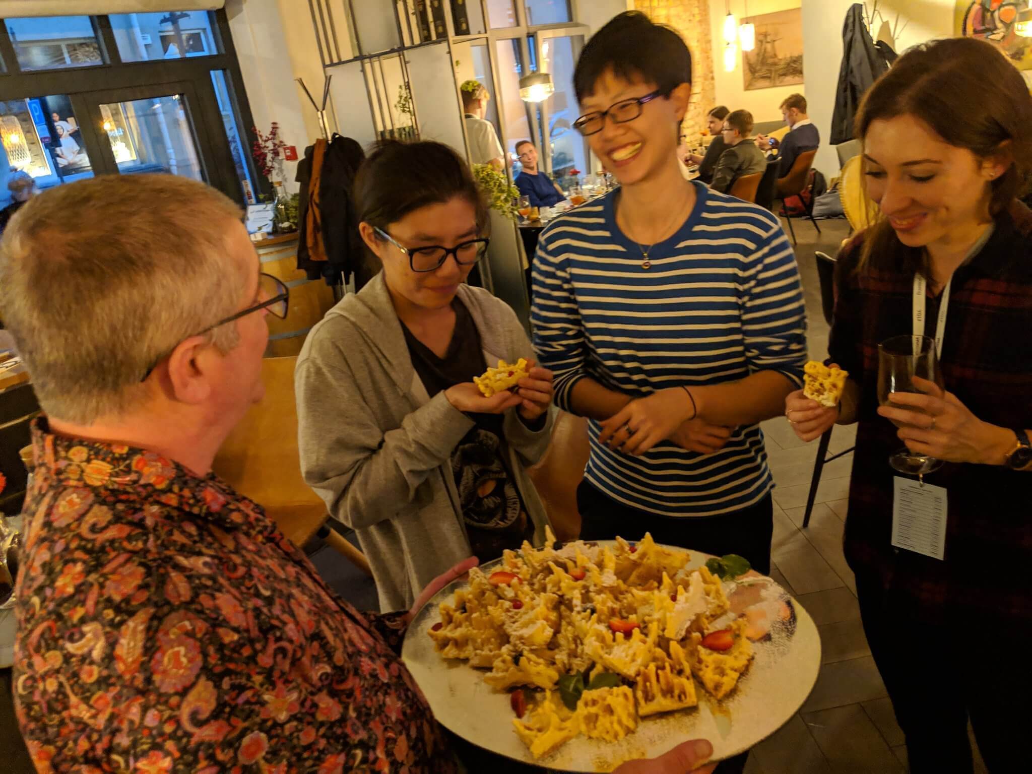 In a restaurant, Bruce is holding a tray of dessert treats in front of the others. He is wearing a lovely / garish paisley shirt.